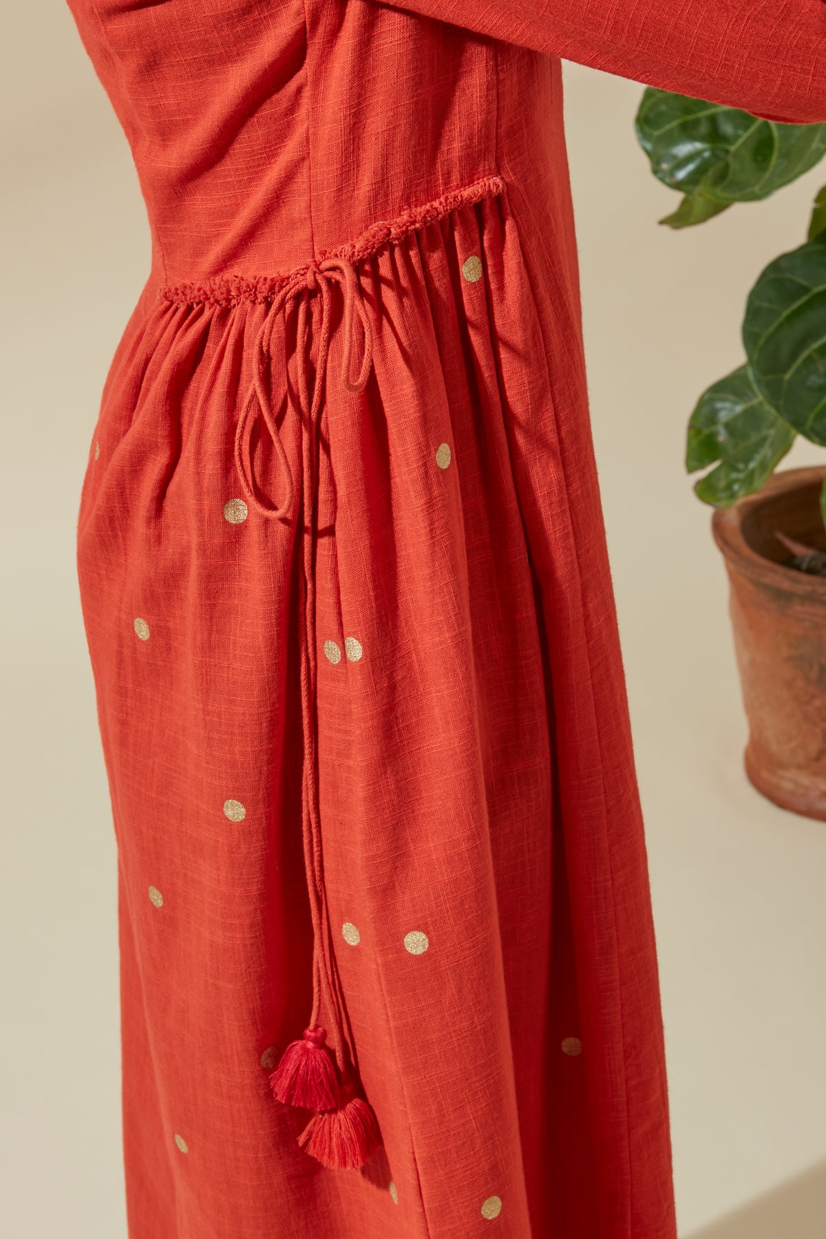 Close-up of Women's Crimson and Gold Cotton Kurta Dress, showing the gathered side tie-up with fringed tassels, fabric texture and gold dot detailing.