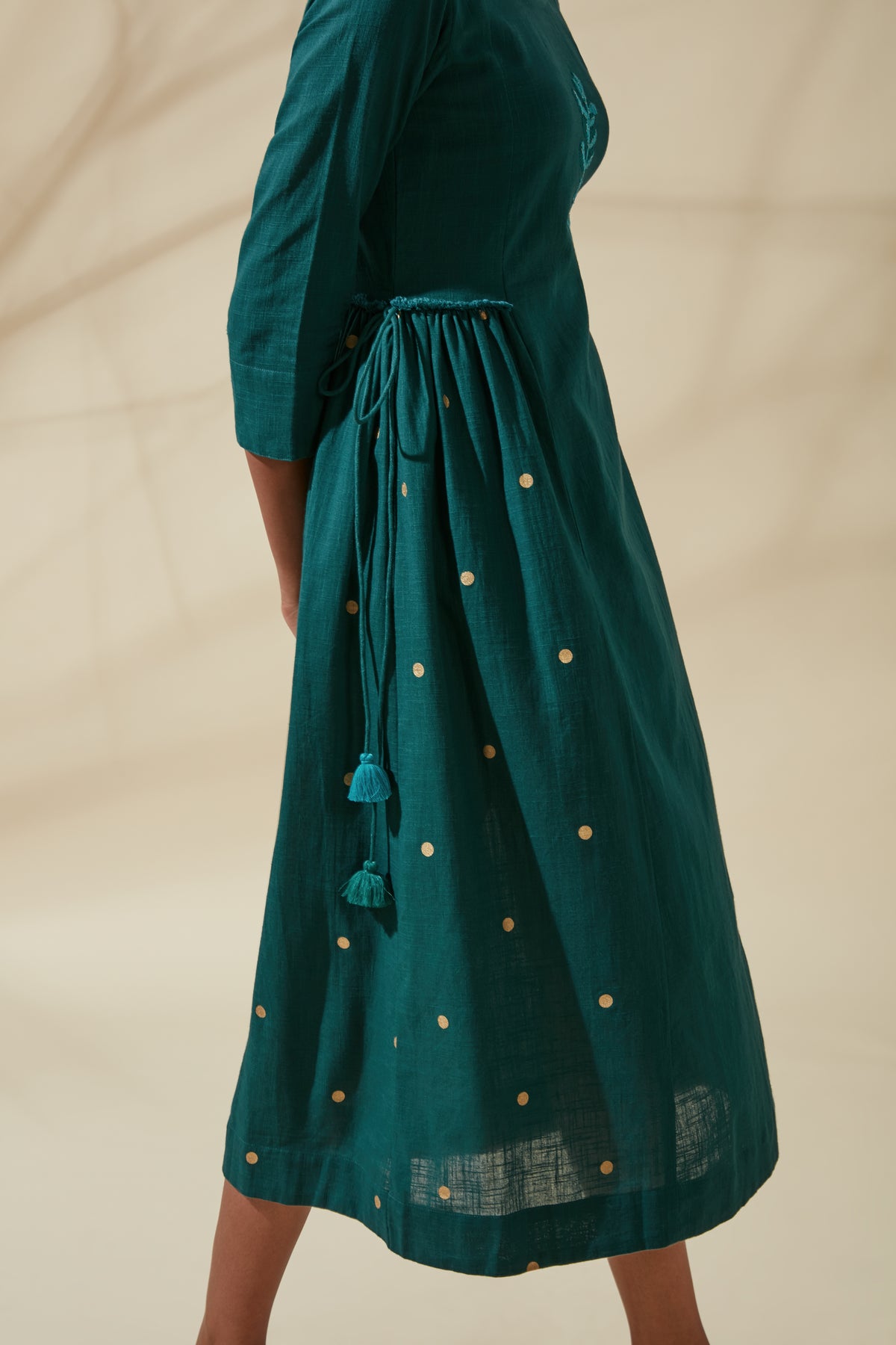 Close-up side view of Women's Teal and Gold Cotton Kurta Dress, showcasing gathered waist ties, hanging tassels and gold dotted accents on the flared skirt.